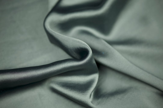 Nahaufnahme eines luxuriösen Satinstoffs in elegantem Salbeigrün mit sanftem Schimmer und natürlichem Faltenwurf – ideal für dezente Mode und Wohntextilien.